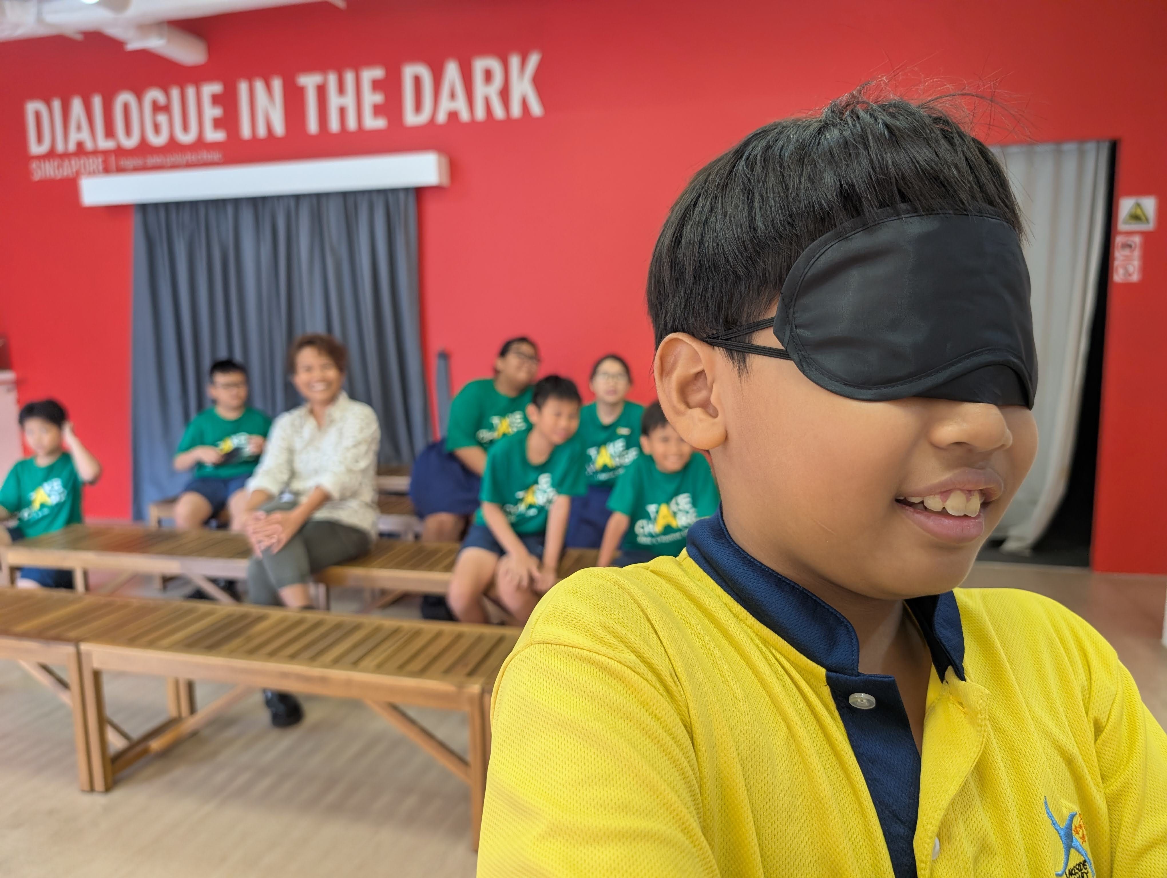 A student wearing a black blindfold attempts to draw as fellow students and their teacher watches on from the back